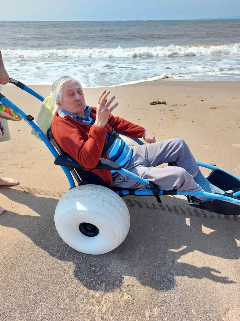Edward having a great time outside Edward Perryman in a beach wheelchair waving happily.