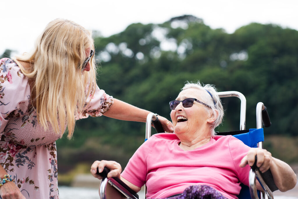 Residential Dementia Care - Lisa Burge from the Sefton Hall care team at the beach in Dawlish with resident Shirley Mapston. Smiling and enjoying the outdoors.