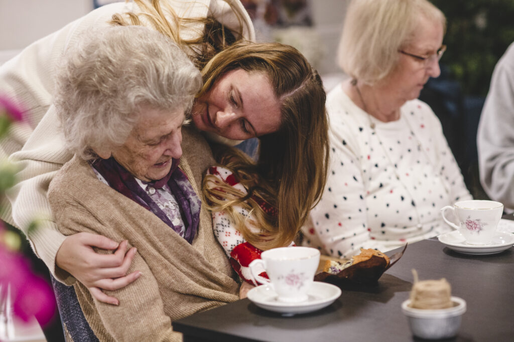 A team member giving a resident a hug around the shoulders whilst they sit and have tea.