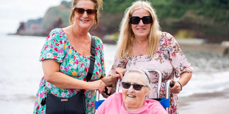 Julie-Yates-and-Lisa-Burge-from-the-Sefton-Hall-care-team-at-the-beach-in-Dawlish-with-resident-Shirley-Mapston who is in a beach wheelchair