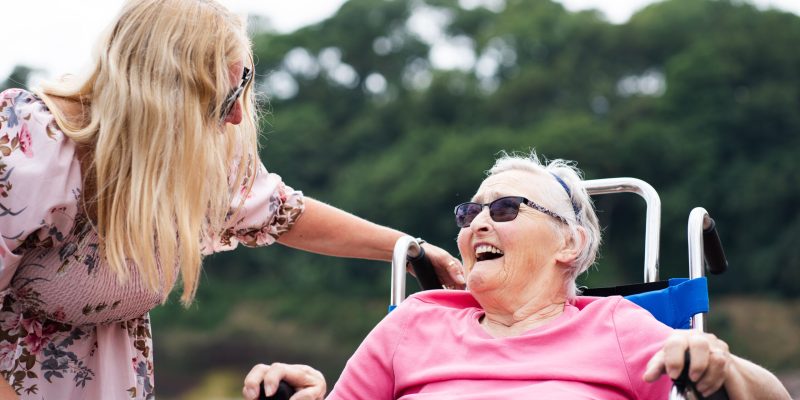 Residential Dementia Care - Lisa Burge from the Sefton Hall care team at the beach in Dawlish with resident Shirley Mapston. Smiling and enjoying the outdoors.